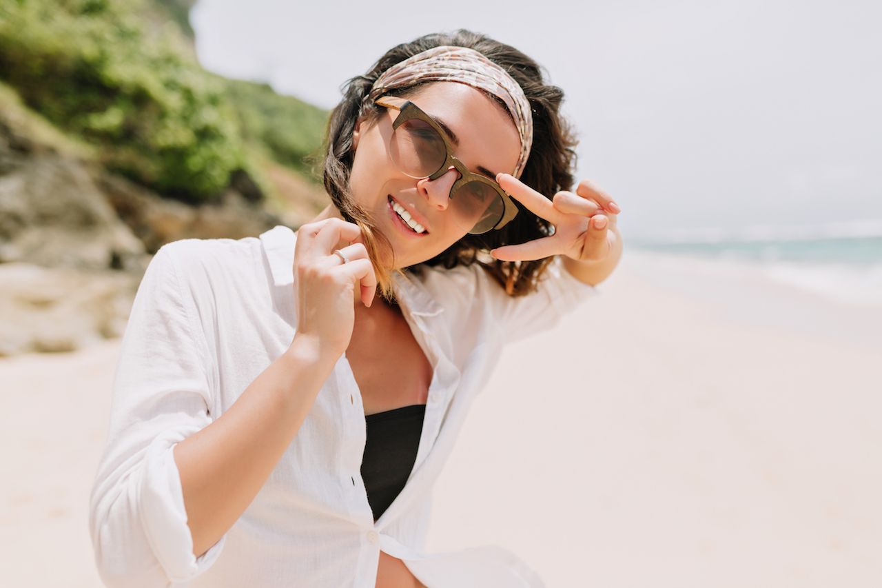 happy-exited-young-woman-with-dark-wavy-hair-wears-black-glasses-hair-ornament-white-shirt-walks-down-white-sandy-beach-sunlight.jpg